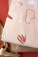 Tissu Sandstorm rose poudré à motif de formes abstraites contours terracotta et écru, plié sur un support en bois devant un fond terracotta.