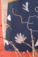 Tissu Sandstorm marine à motif de formes abstraites contours écru et moutarde, plié sur un support en bois devant un fond terracotta, avec des brindilles séchées dorées.
