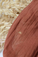 Sunset fabric in terracotta, a semi-sheer cotton gauze with sun motifs embroidered in gold thread, placed near a palm leaf.