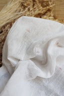 Tissu Sunset écru en gaze de coton semi-transparente, brodé de petits motifs soleil terracotta, posé sur un tapis en fibres naturelles près d'épis de blé séchés.