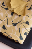 Feuille de ginkgo dorée posée sur le tissu Windy moutarde plié, à motif de feuilles courbes marine et écru.