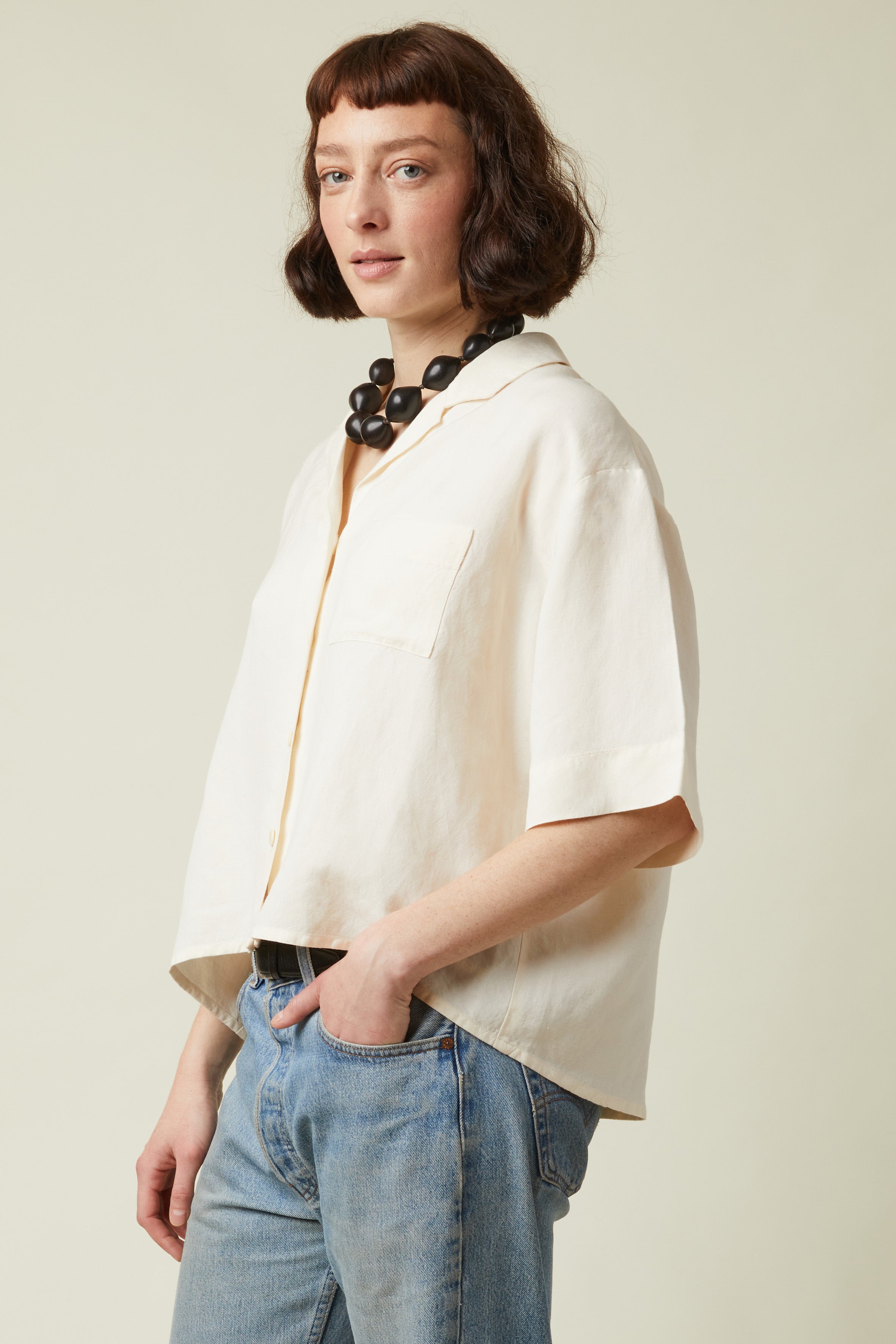 Short-sleeved Shirt pattern – left profile view, short-sleeved shirt crème blouse collar, pearl necklace, jeans – Atelier Brunette