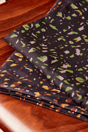 Gros plan sur le tissu viscose imprimé confettis façon terrazzo anthracite, à éclats verts, lavande, orange et écru, plié sur une table en bois.