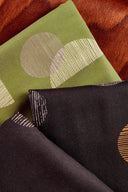 Gros plan sur les tissus viscose Jane pliés, noir et vert matcha, motifs graphiques ronds à rayures beige et orange, posés sur un fond cuir cognac.