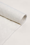 Le Coton Indien Tissé 001 ecru fabric rolled up, with the raised woven floral pattern visible along its full length and the frayed selvedge showing.